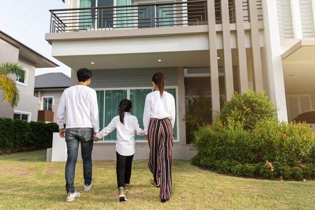 Parents and child walk hand in hand across a lawn toward a modern two-story house, seen from behind.