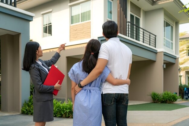 Real estate agent points to a modern house while a couple stands arm in arm, viewing the property exterior.