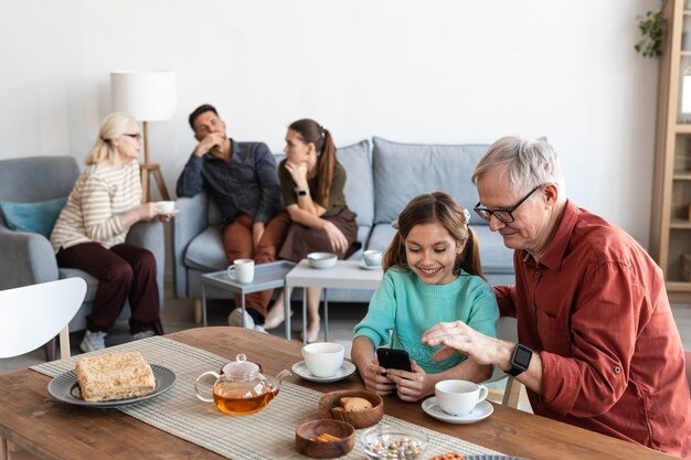 An elderly man is showing a smartphone to his granddaughter at a cozy tea table while other family members talk on the sofa.