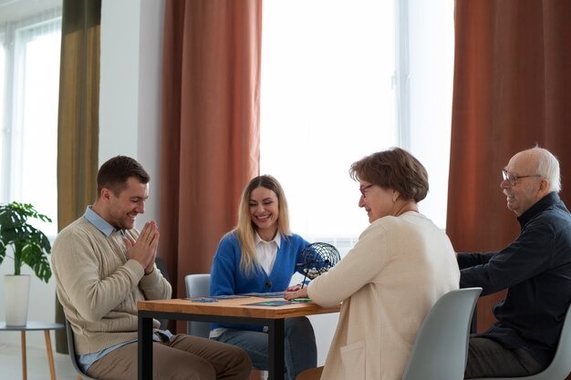Seniors and a younger caregiver are sitting around a table playing bingo together in a bright community room.