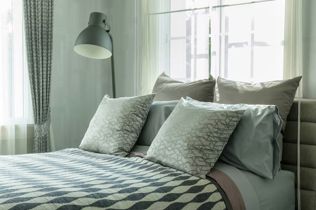 Bright bedroom with patterned pillows, geometric blanket, and a floor lamp beside a window.

